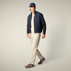 A man with dark hair faces away from the camera, wearing the SAVE THE DUCK - SEDUM JACKET in dark navy by SAVE THE DUCK, paired with light beige pants and brown shoes, set against a plain light background.