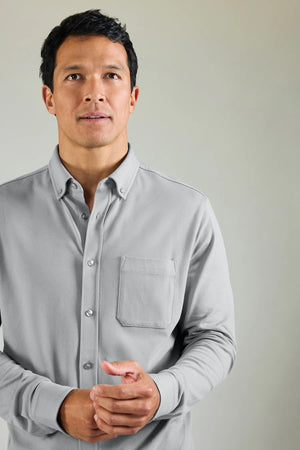 A man with short dark hair wears the MACK WELDON Silver Pique Long Sleeve Button Up Shirt in True Navy, standing against a plain light background with a neutral expression and his hands together in front of him.