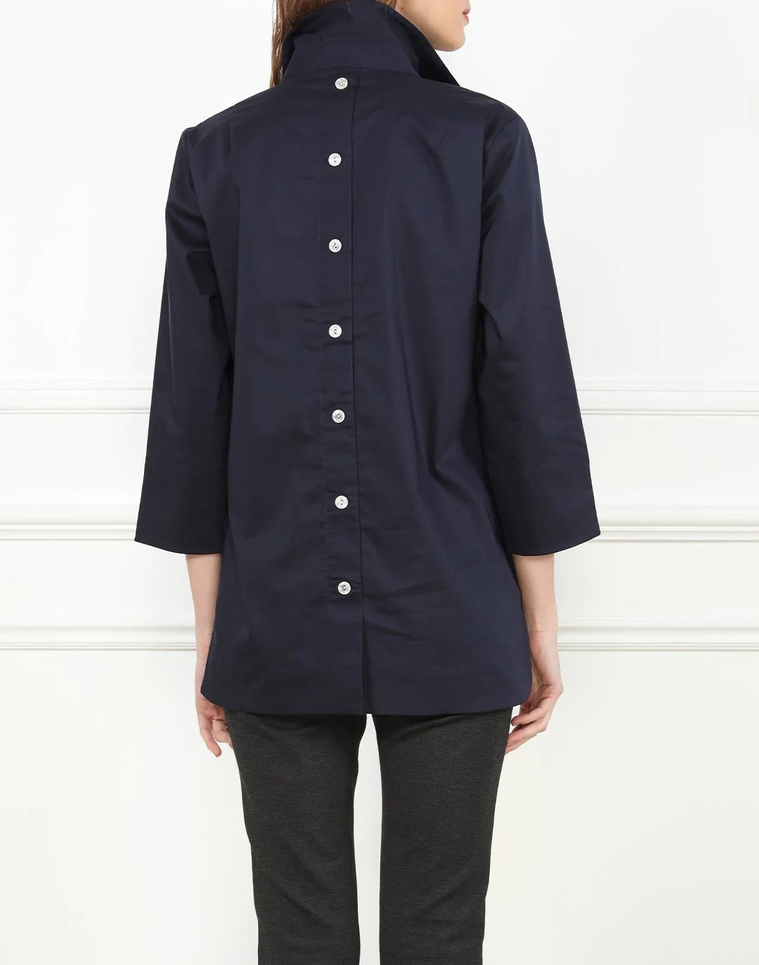 Sporting long hair, the individual is clad in a white HINSON WU - AILEEN TUNIC and black pants. The tunic-length shirt by Hinson Wu features a collar and two buttons near the top. They stand in front of a light gray wall with white molding, with their face not visible in the image.