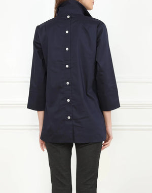 Sporting long hair, the individual is clad in a white HINSON WU - AILEEN TUNIC and black pants. The tunic-length shirt by Hinson Wu features a collar and two buttons near the top. They stand in front of a light gray wall with white molding, with their face not visible in the image.