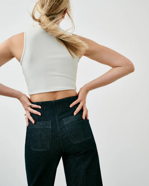 A woman is seen from behind, wearing a white sleeveless crop top and dark high-waisted SPANX - EVERYWEAR CROPPED WIDE LEG DENIM. Her hands are placed on her hips, with her blonde hair falling over one shoulder. The plain light gray background perfectly highlights her stylish SPANX denim that offers impeccable shape.
