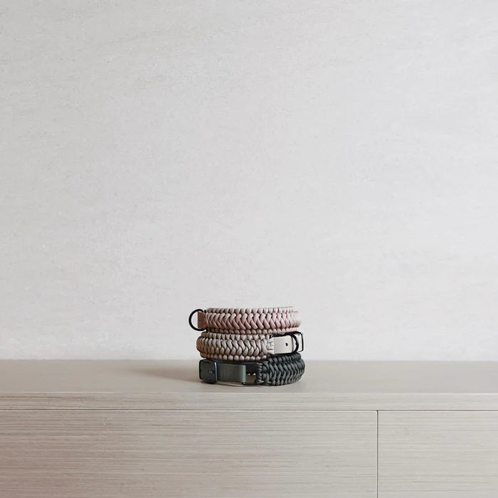 Two LAMBWOLF COLLECTIVE NEW JUPITER Dog Collars in small, one in beige and one in gray, are stacked on a light wooden surface against a plain, light-colored wall. Designed by LAMBWOLF COLLECTIVE, each collar combines cushioned comfort with sturdy durability, making them perfect for everyday wear.