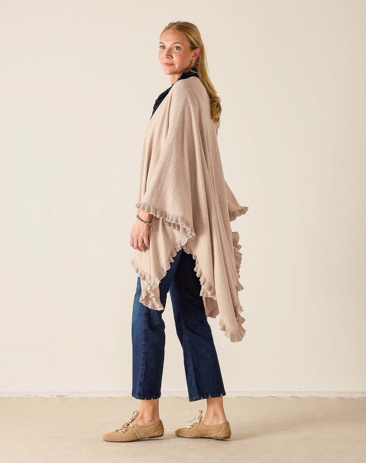 Wearing the MERSEA Charleston Ruffle Wrap in Mink over a white blouse and jeans, a woman leans against a wooden storefront, holding a magazine and gazing to the side.