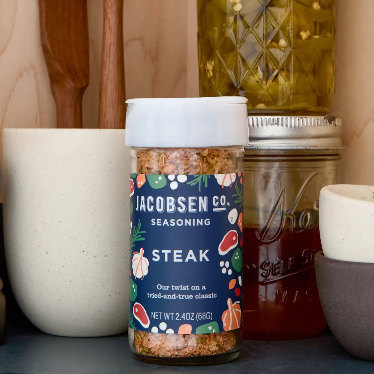 A 2.4 oz (68g) jar of JACOBSEN SALT CO. JACOBSEN SALT - STEAK SEASONING features a blue label decorated with illustrations of garlic, herbs, and meat. Expertly crafted with kosher salt, this seasoning blend is perfect for enhancing your favorite steak cuts.