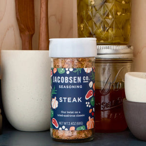 A 2.4 oz (68g) jar of JACOBSEN SALT CO. JACOBSEN SALT - STEAK SEASONING features a blue label decorated with illustrations of garlic, herbs, and meat. Expertly crafted with kosher salt, this seasoning blend is perfect for enhancing your favorite steak cuts.