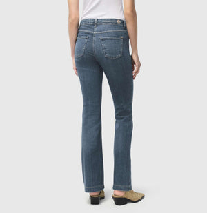 A person stands wearing a blue sweater, white shirt with a bow, and MAC's DREAM BOOT AUTHENTIC DENIM jeans. They have short light brown hair and pose against a plain light gray background.