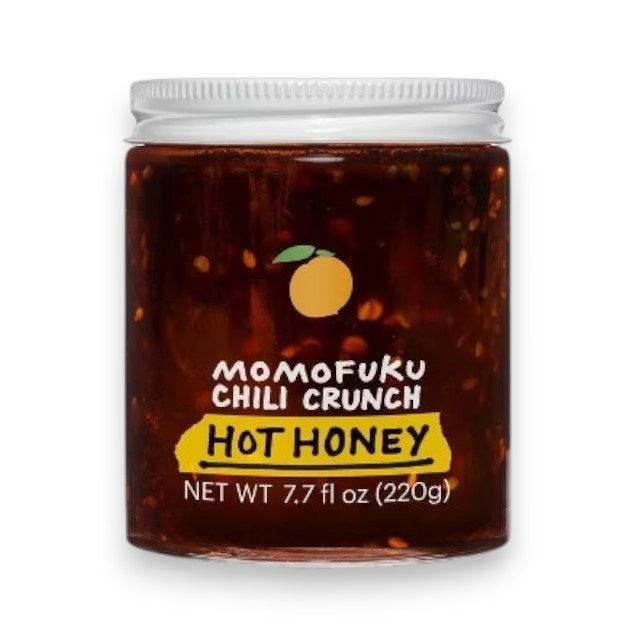 A jar of MOMOFUKU - CHILI CRUNCH HOT HONEY, with a white lid, contains a red-brown mixture inside. The label features an illustration of a citrus fruit along with the text "Momofuku Chili Crunch Hot Honey" and "Net Wt 7.7 oz (220g)." This spicy-crunchy chili oil offers the perfect blend of heat and sweetness.