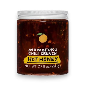 A jar of MOMOFUKU - CHILI CRUNCH HOT HONEY, with a white lid, contains a red-brown mixture inside. The label features an illustration of a citrus fruit along with the text "Momofuku Chili Crunch Hot Honey" and "Net Wt 7.7 oz (220g)." This spicy-crunchy chili oil offers the perfect blend of heat and sweetness.