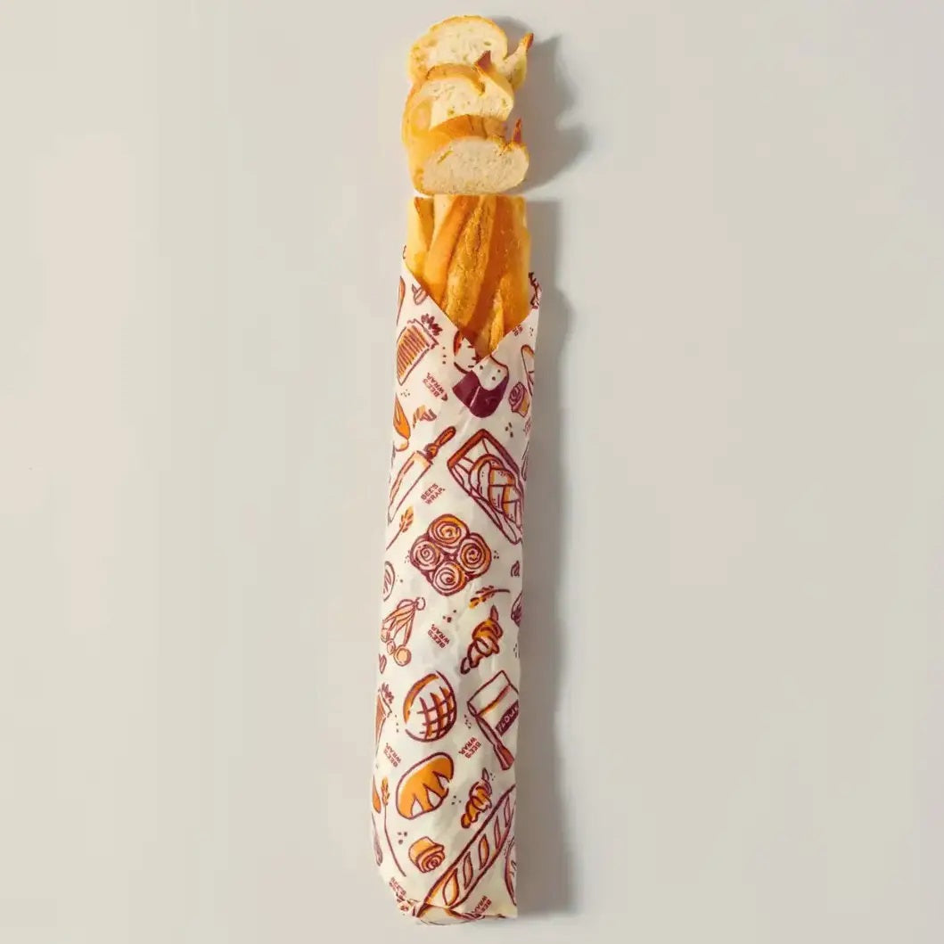 A loaf of bread is partially tucked inside the BEES WRAP- BEESWAX REUSABLE BREAD BAKERY LANE bag by BEES WRAP, featuring a small white label and displayed against a plain white background—a perfect choice for a plastic-free kitchen.