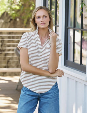 A woman with blonde hair stands outdoors, looking at the camera, wearing the FRANK & EILEEN Charlotte Perfect Polo in Heritage Jersey Wintage White and Camel Stripe. Greenery and white flowers are softly blurred in the background.