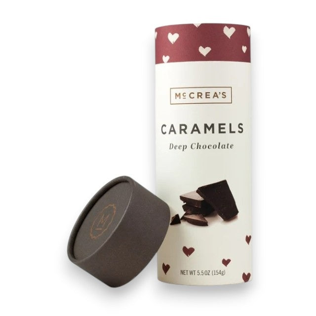 A cylindrical container of McCREA’S CANDIES - DEEP CHOCOLATE CARAMELS decorated with heart designs, shown with the lid off and resting nearby. The label features images of chocolate pieces, making it an ideal choice for anyone who enjoys creamy, fresh milk chocolate caramels from McCREA’S CANDIES.
