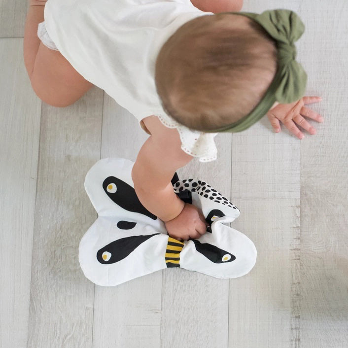 The WEE GALLERY - BUTTERFLY ORGANIC CRINKLE is a children's soft fabric toy, crafted from organic cotton. It features a butterfly shape with a smiling face in the center and a striped yellow and black body. The white wings are adorned with black patterns and yellow accents, providing visual stimulation. Additionally, it includes a small loop at the top for hanging.