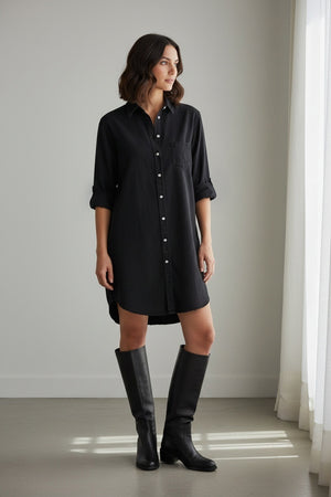 A woman in the FRANK & EILEEN Mary Classic Shirtdress in Famous Denim Black stands outdoors on a stone path, smiling with one hand on her hip. She wears a wide-brimmed straw hat; green plants and a large window are in the background.