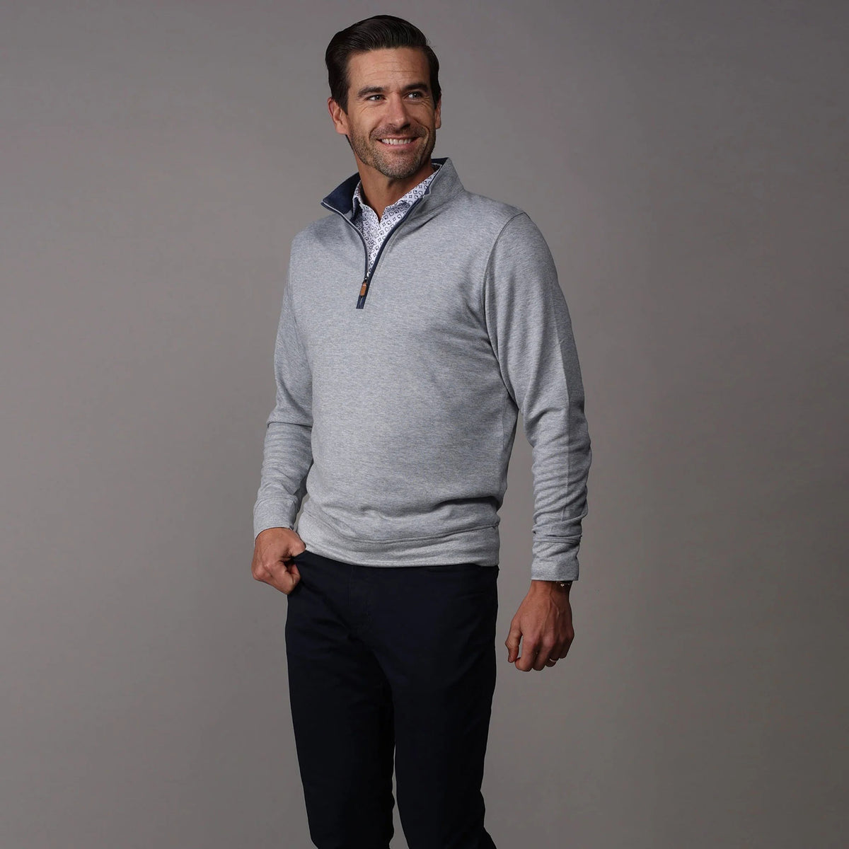 A man with short dark hair and a beard smiles, wearing the COLLARS AND CO - CLOUD COTTON QUARTER ZIP PULLOVER IN HEATHER GREY over a patterned shirt. He stands against a plain gray background with his hands in his pockets.