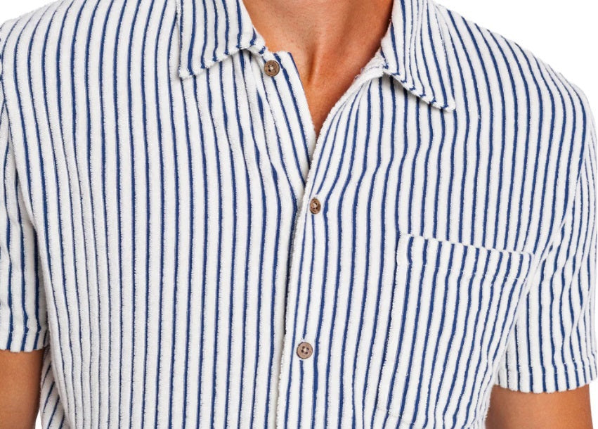 A man with curly hair and a short beard wears the CRIQUET Terrycloth Button Down Shirt in Striped Jacquard Natural, holding a drink in a koozie. Surfboards, sand, and trees create a relaxed summer vibe in the background.