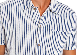 A man with curly hair and a short beard wears the CRIQUET Terrycloth Button Down Shirt in Striped Jacquard Natural, holding a drink in a koozie. Surfboards, sand, and trees create a relaxed summer vibe in the background.