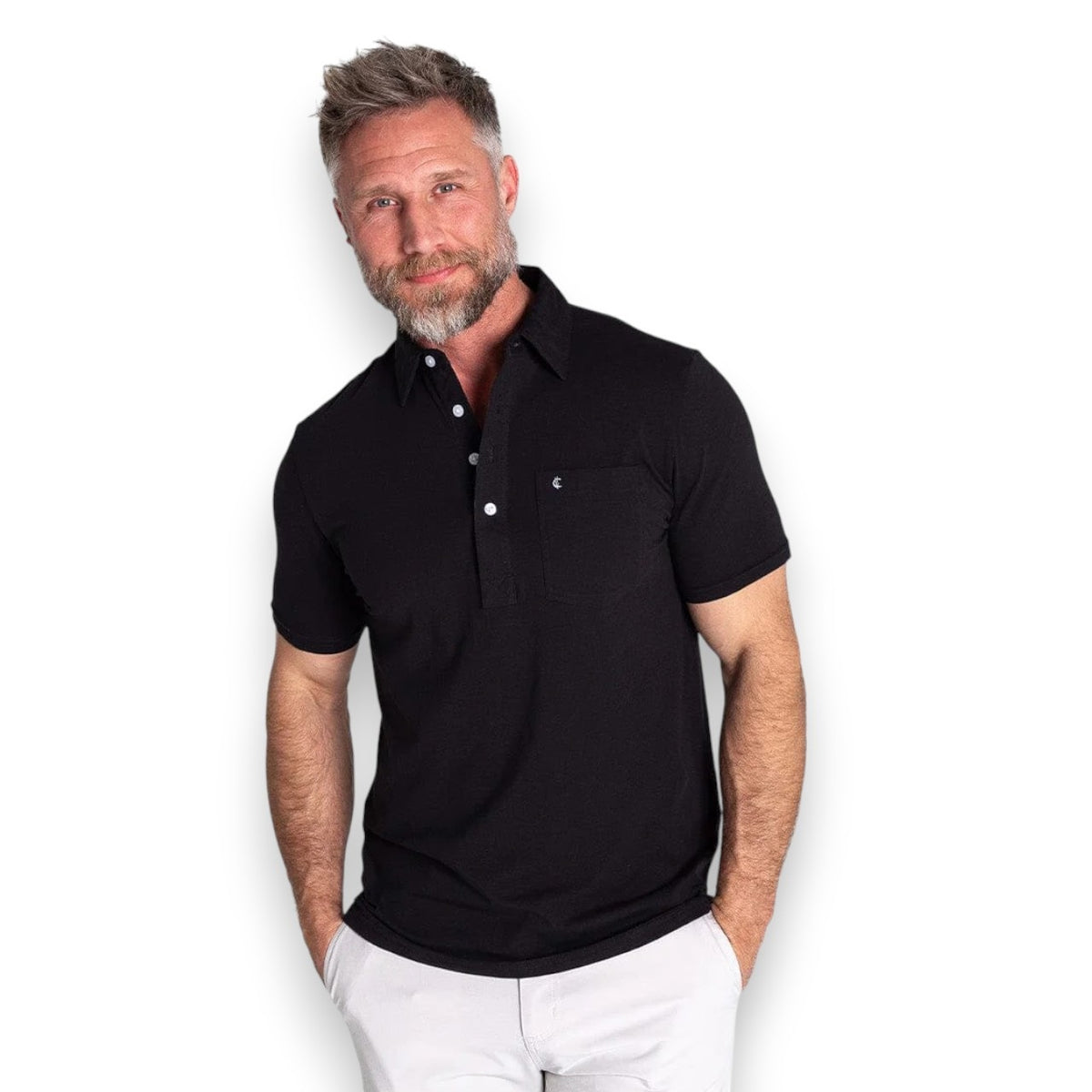 A man with short gray hair and a beard wears the CRIQUET Top Shelf Players Shirt in Black by CRIQUET, paired with light-colored pants, standing with hands in his pockets against a plain white background.