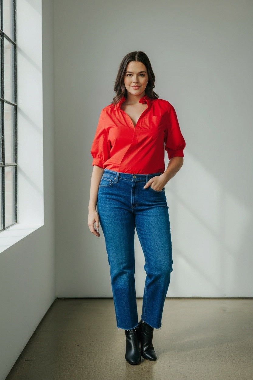A woman in the KMJ - Southern Lover Shirt with a ruffle collar and blue jeans, featuring the iconic KMJ tuck, stands confidently beside a horse, holding its reins amidst green foliage. She's elegantly posed with jewelry, embodying Southern-inspired charm.