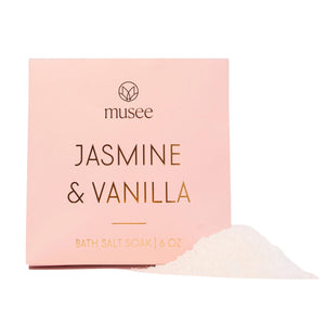 A pink packet labeled “MUSEE - JASMINE & VANILLA MINI BATH SALT SOAK” stands upright on a light background with a slight shadow to the right. Infused with calming jasmine and vanilla along with relaxing Pacific sea salt, this bath blend from MUSEE offers a soothing, spa-like experience at home.