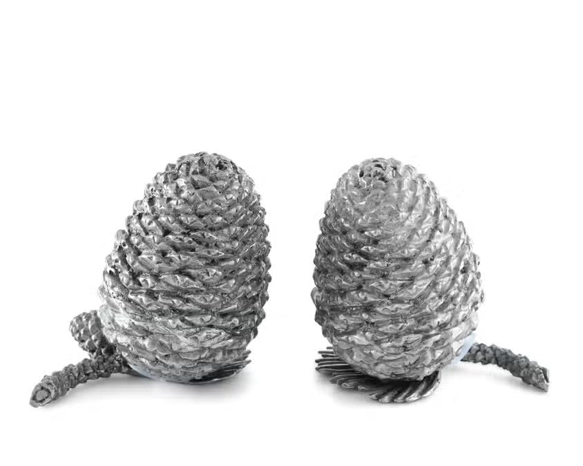 Two detailed silver pine cones stand upright on a white background, resembling the VAGABOND HOUSE - PEWTER PINE CONESALT AND PEPPER SET by VAGABOND HOUSE, with small pine needle segments visible at their bases.