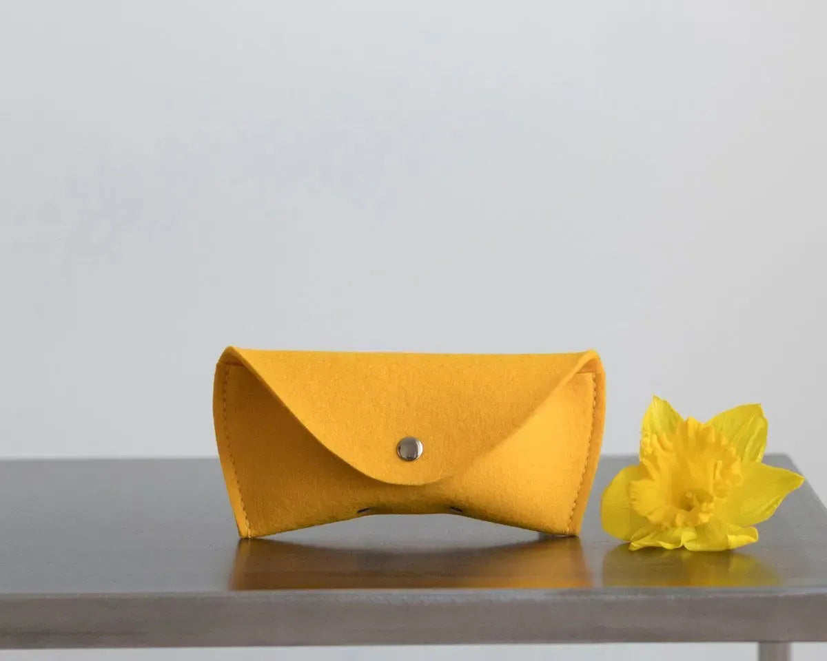 Three WOOLBERRY WOOL FELT GLASSES CASES in navy, bright, and light blue with magnetic closures are arranged on a white surface near clear sunglasses, framed by white-blue hydrangeas.