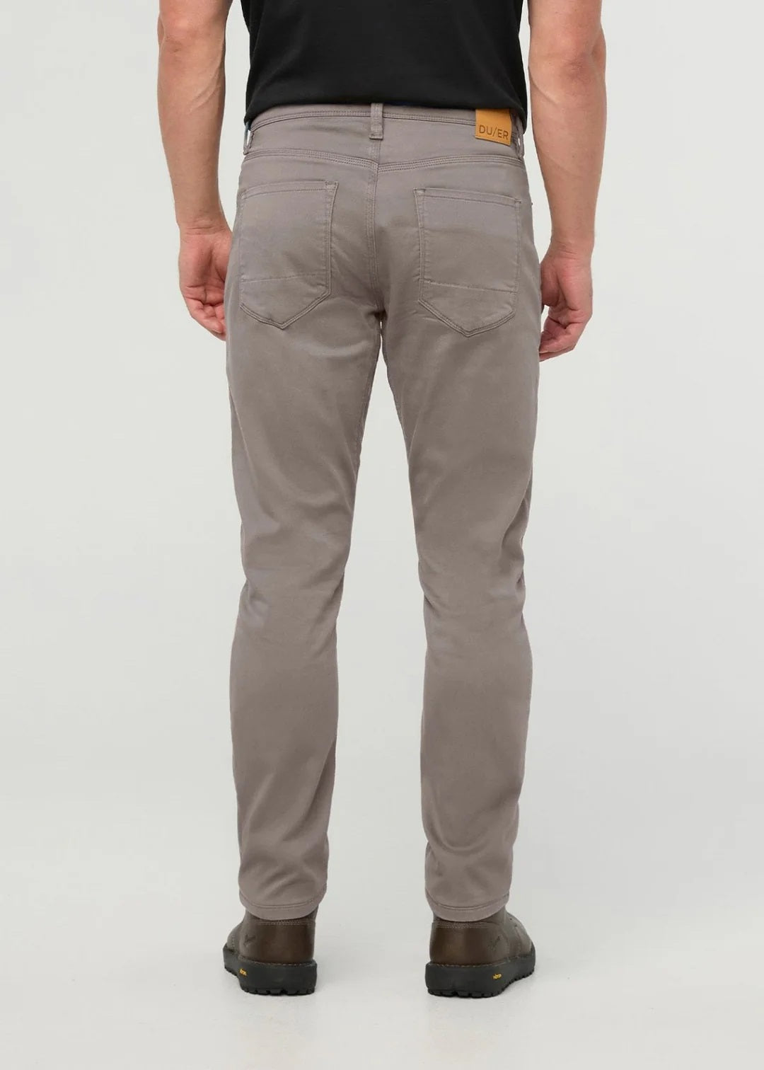 Close-up of the back of a person wearing DUER No Sweat Athletic Taper Pants in Stone, featuring two back pockets and a tan leather DUER patch on the waistband. They're also wearing a black shirt.