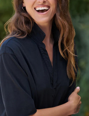 A woman in a loose, short-sleeve FRANK & EILEEN MINI EMMA Popover Henley Dress in Vintage Jersey British Royal Navy from the California Heritage Collection stands on a gravel path, smiling and looking down while holding a white wide-brimmed hat. Lush green foliage lines the background.