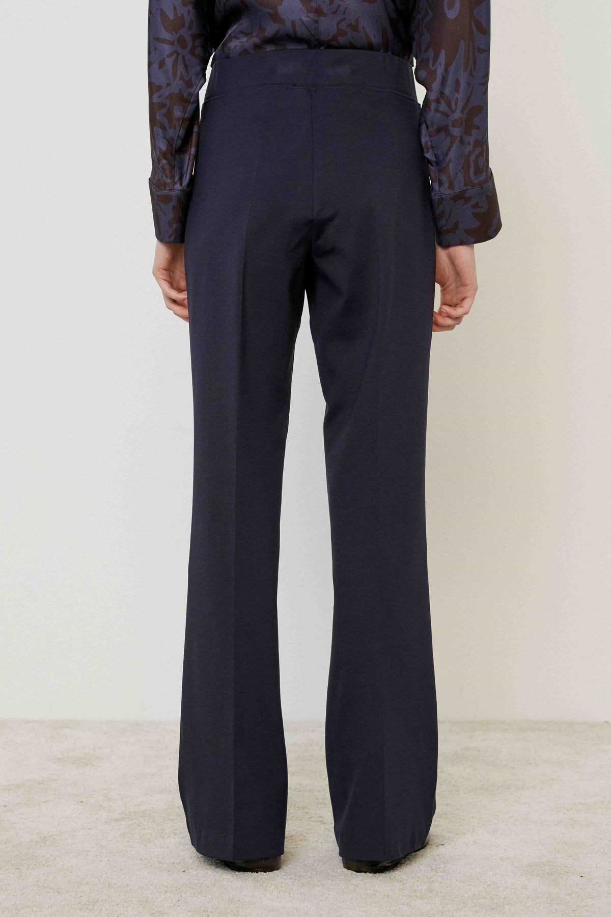 A person in MEIMEIJ PULL ON PONTE TROUSER PANT, a patterned long-sleeve blouse, and black shoes stands on a light carpet against a plain wall, highlighting this versatile MEIMEIJ wardrobe essential with a tailored look.