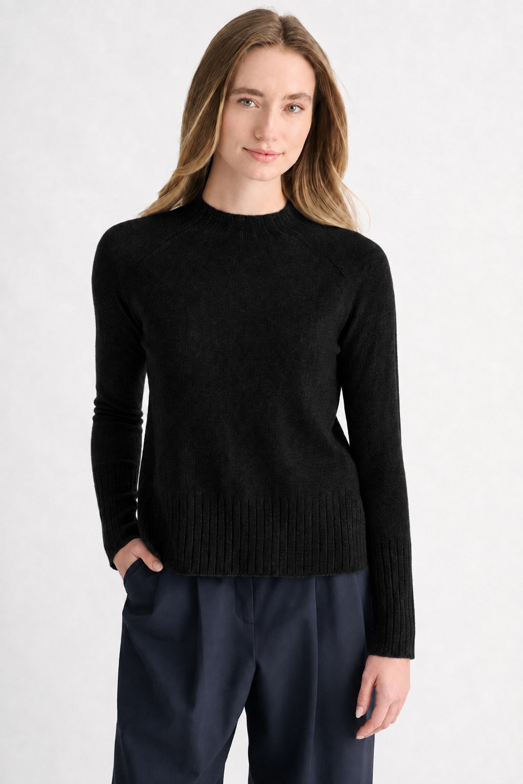 A person with long light brown hair is wearing the KINROSS CASHMERE Hi Low Rib Trim Funnel Cashmere Sweater in Black by KINROSS, paired with dark pants. They stand against a light background with one hand in their pocket, looking at the camera and smiling slightly.