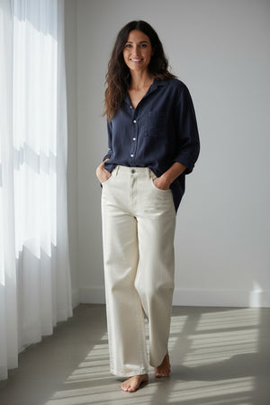 A woman stands barefoot indoors, smiling with her hands in her pockets, dressed in the FRANK & EILEEN EILEEN Relaxed Button Up Shirt in Italian Cashmere Touch Navy and cream wide-leg pants. Sunlight streams through sheer curtains, casting soft shadows across the floor.