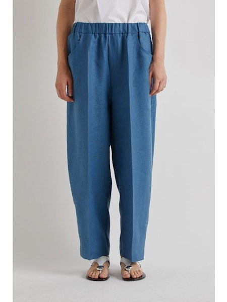 A person stands against a plain light background, cropped at the shoulders, wearing MEIMEIJ Pull On Barrel Pant in Cerulean with an elastic waistband, paired with a white shirt and sandals.