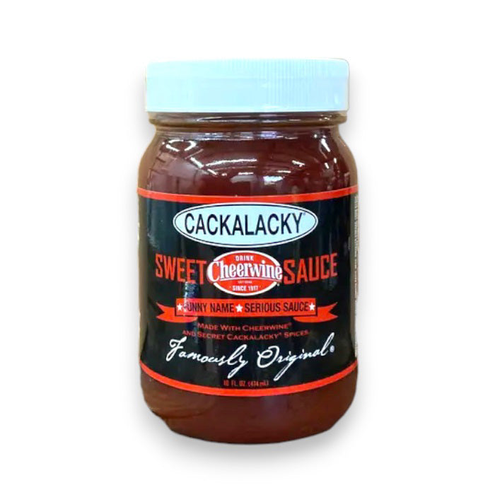 A person brushes savory barbecue sauce onto the burgers sizzling on the grill, holding a jar of CACKALACKY - CHEERWINE SWEET SAUCE in their other hand. This scene of Southern cuisine unfolds against the backdrop of a grassy outdoor setting.