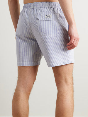 A person wears HARTFORD - SEERSUCKER SWIM TRUNKS IN INDIGO with a drawstring waist, side pockets, and a quick-drying seersucker fabric. The mid-thigh trunks are shown against a plain background, displaying only the torso and legs.