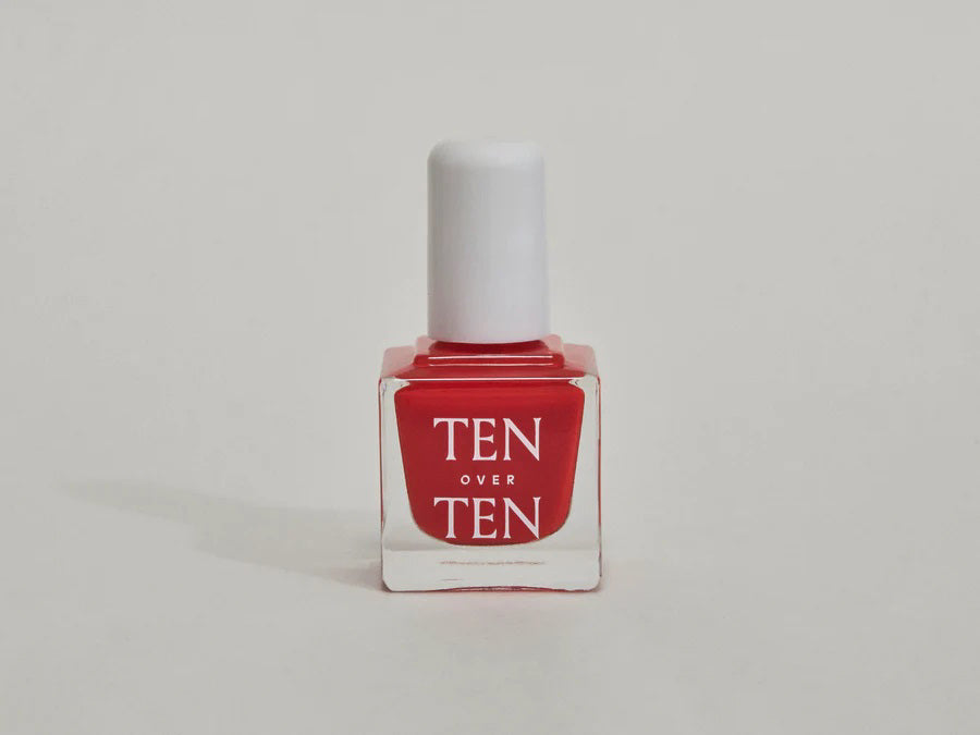 A glass bottle of TENOVERTEN - NAIL POLISH from the brand TEN OVER TEN, featuring a white cap and a dusty pink nail color. The front label displays "TEN OVER TEN" in white uppercase letters. This vegan, eight-free formula ensures both beauty and peace of mind. The background is plain and light-colored.