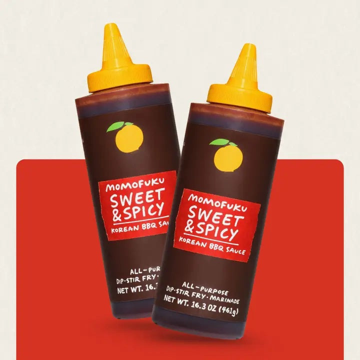 Two bottles of MOMOFUKU - SWEET & SPICY KOREAN BBQ SAUCE from MOMOFUKU, each with a yellow cap and a label featuring a yellow plum graphic, are displayed against a red and beige background—perfect for use as a vegan marinade or for adding bold, gochujang-inspired flavor to your favorite dishes.