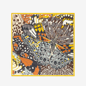 A close-up of a person wearing the INOUI EDITIONS - PAPILLON (BUTTERFLIES) SQUARE WOOL SCARF IN NATURAL by INOUI, draped around their neck over a black top. The scarf showcases abstract and geometric butterfly designs in earthy tones.