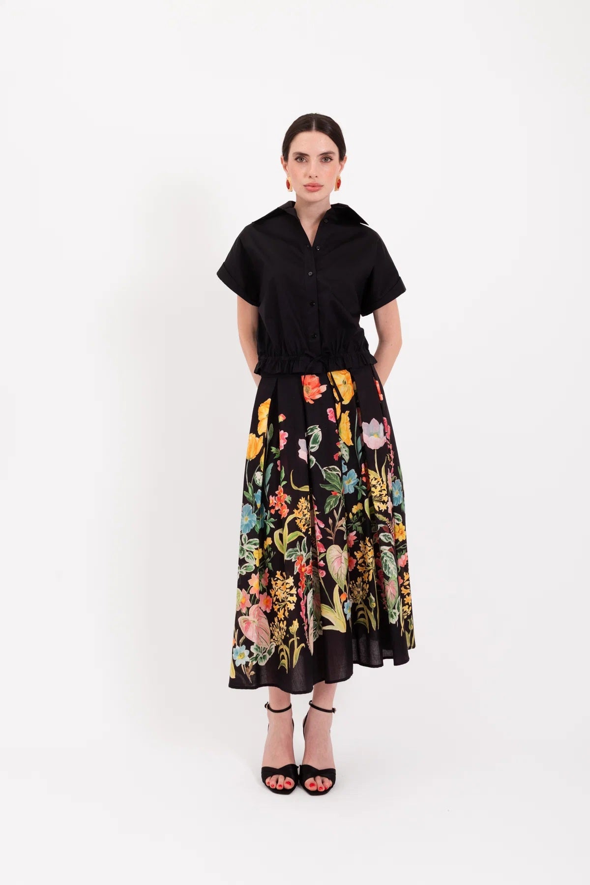 A woman stands against a white background wearing a black short-sleeve shirt paired with the LAVI - IRIS Botanical Skirt in Black Floral from LAVI, featuring colorful floral patterns, and completes the look with black high-heeled sandals.