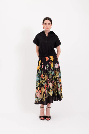 A woman stands against a white background wearing a black short-sleeve shirt paired with the LAVI - IRIS Botanical Skirt in Black Floral from LAVI, featuring colorful floral patterns, and completes the look with black high-heeled sandals.