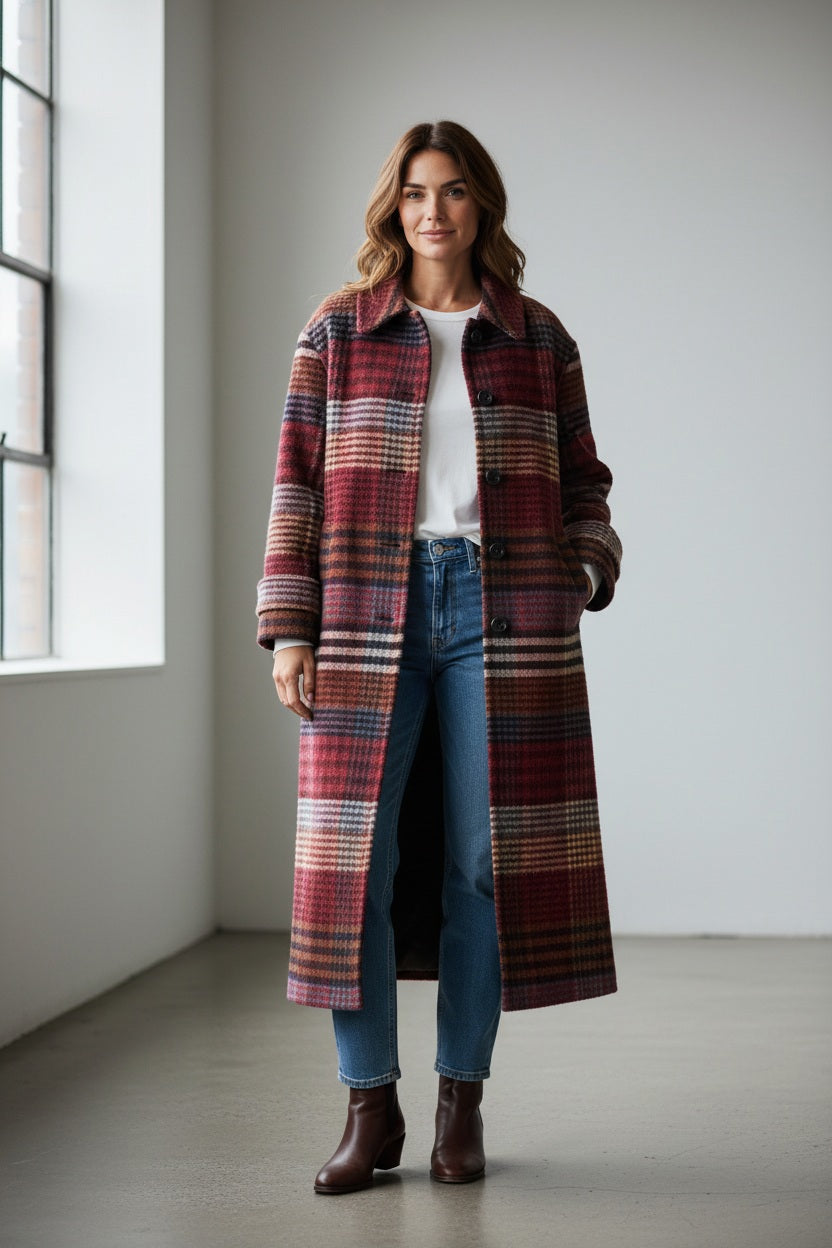 A woman stands indoors on light wooden flooring, barefoot and bathed in natural window light, effortlessly dressed for cold weather in the MIRTO 1956 - LONG PLAID WOOL COAT layered over a floral bow-tie blouse and a long burgundy skirt.