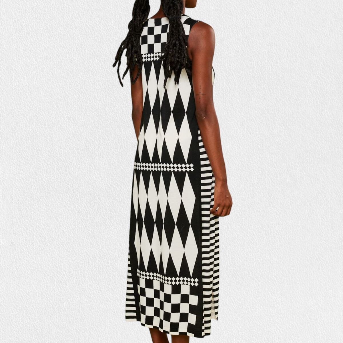 A person is wearing the IN BED WITH YOU - LONG KNIT DRESS featuring a sleeveless, knee-length design and a striking geometric pattern. The dress, from the Italian brand IN BED WITH YOU by CLAUDIOLAI SRL, showcases alternating squares and circles in vibrant prints of red, yellow, purple, and white set against a plain white textured background.