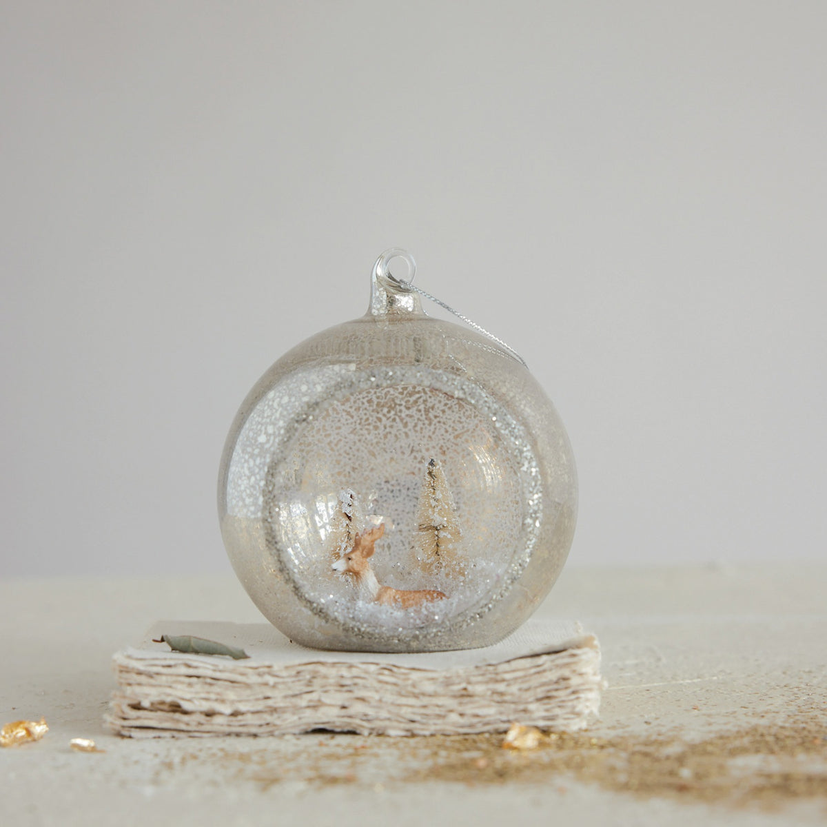 A MERCURY GLASS DIORAMA ORNAMENT WITH DEER, BOTTLE BRUSH TREES AND GLITTER featuring a miniature deer and bottle brush tree inside a mercury glass globe with a snowy diorama.
