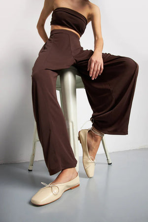 The Augusta - Sienna Ballet Flat by AUGUSTA is a single silver-colored shoe featuring a subtle metallic sheen and a simple bow on the front. It has a low brown sole and a pull tab at the back. Handcrafted in Spain from leather, this ballet flat is shown from the side against a light gray background.