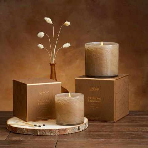 A candle in a transparent glass container, featuring a rich brown hue, is placed beside a coordinating brown box adorned with gold details. The set exudes a gourmand aroma and is elegantly labeled as &quot;LAFCO - Pumpkin Seed &amp; Sandalwood Candle 6.5oz.&quot; The candle&#39;s texture appears soft like fur.
