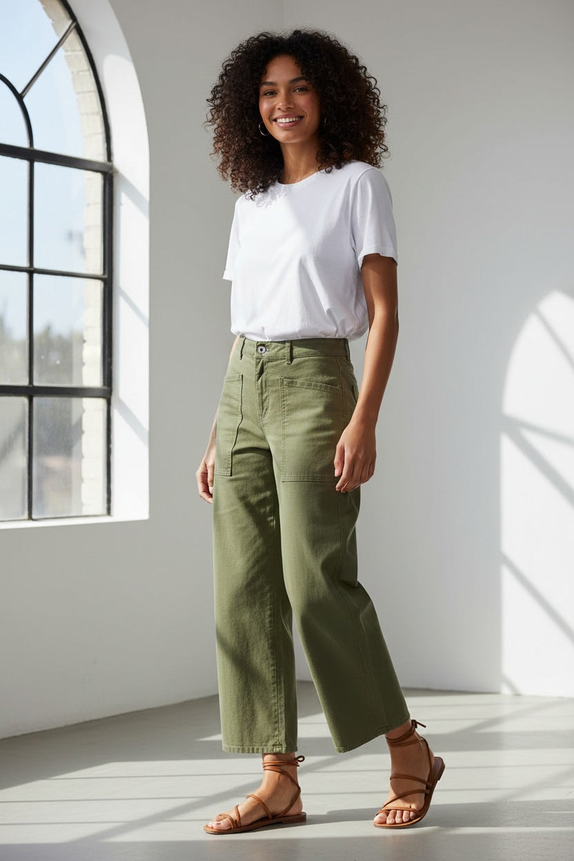 Outdoors on grass, a person wears the FRANK &amp; EILEEN Bray Everyday Barrel Pant in Italian Peached Twill Olive with large front pockets and a white top. One hand is in a pocket, with greenery blurred in the background.