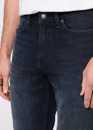 A man stands against a plain background wearing a black jacket, white t-shirt, DU/ER - PERFORMANCE DENIM+ SLIM STRIAGHT PANTS IN CARBON STONE, and black shoes, with one hand in his pocket and a neutral expression.