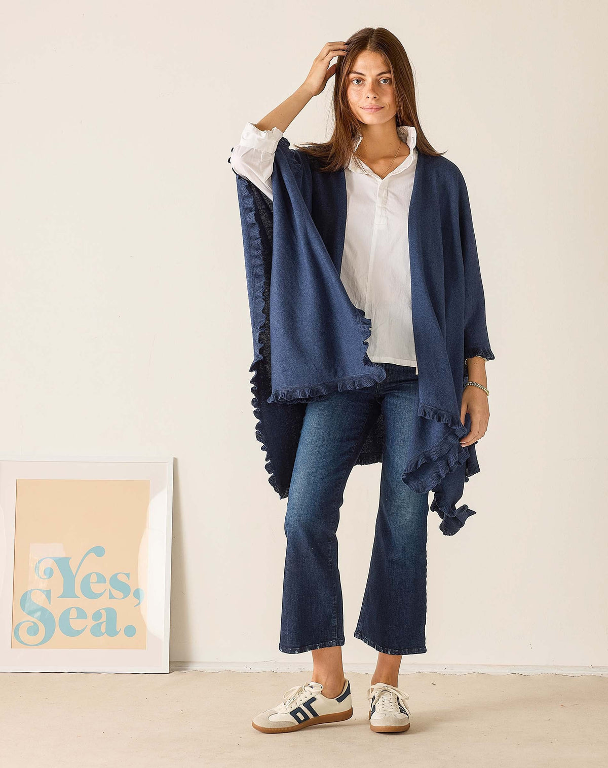 A woman with long brown hair sits sideways on a wooden chair, wearing the MERSEA - CHARLESTON RUFFLE WRAP IN CHAMBRAY over a white shirt, jeans, and sneakers, looking at the camera against a plain background.