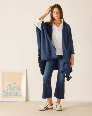 A woman with long brown hair sits sideways on a wooden chair, wearing the MERSEA - CHARLESTON RUFFLE WRAP IN CHAMBRAY over a white shirt, jeans, and sneakers, looking at the camera against a plain background.