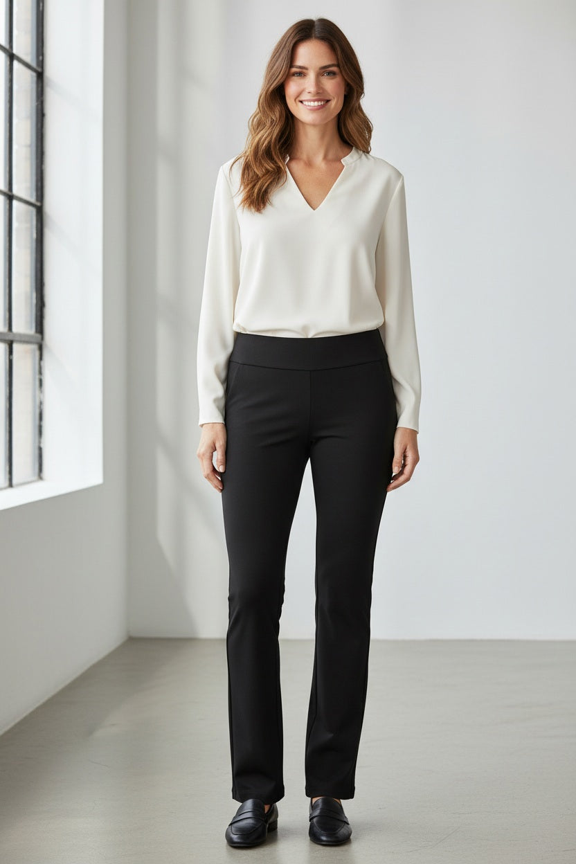 A woman stands indoors wearing a white long-sleeve blouse and the PULL ON STRIGHT LEG MICROFIBER PANT - LONG by KRAZY LARRY, INC, featuring a 32″ inseam in black. She pairs the outfit with black shoes and smiles while facing forward in a bright, minimalistic room with large windows.