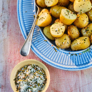 A box of GOURMET FOOD SOLUTIONS' MALDON - GARLIC SEA SALT is placed on a bed of coarse salt with herbs and garlic flakes scattered around. The box, in a green and white geometric design, reads "MALDON - GARLIC SEA SALT" and "with white & roasted garlic," established 1882—proudly showcasing its English sea salt heritage.