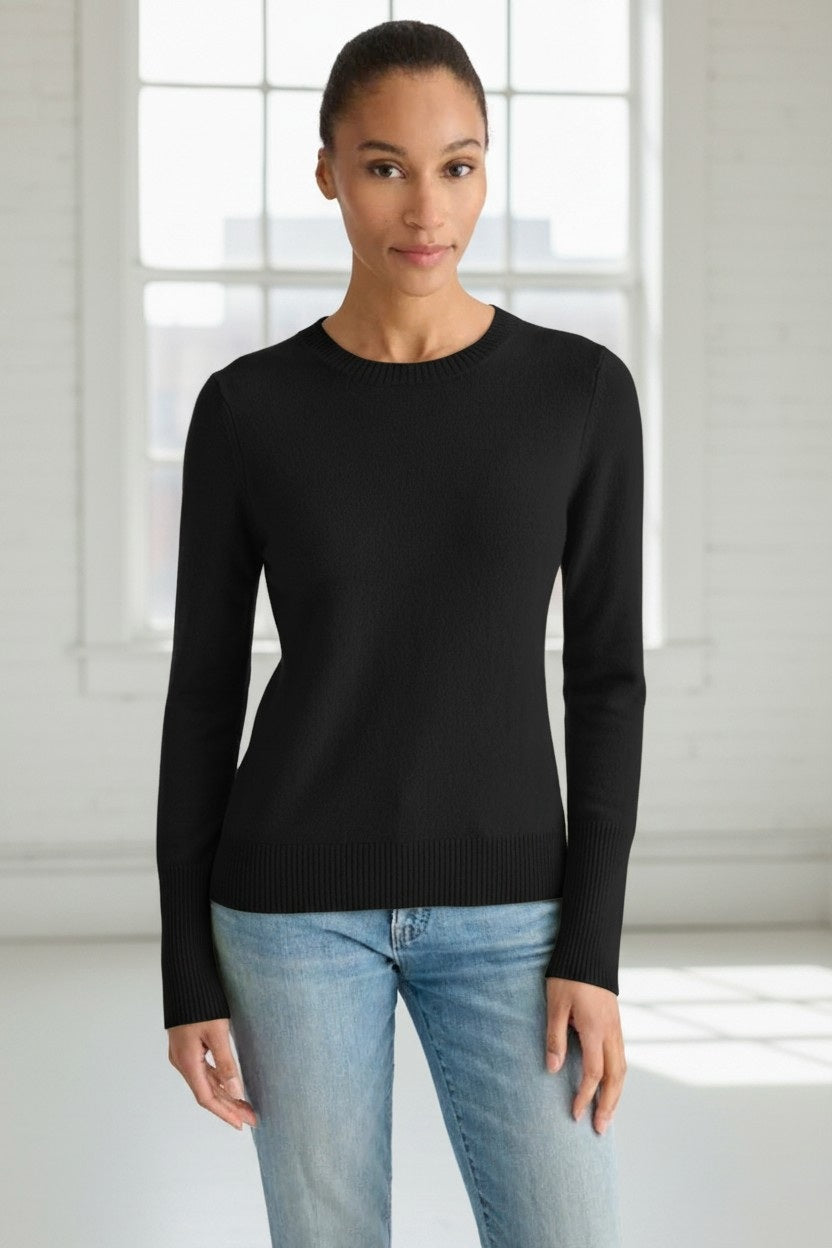 A woman stands indoors in a bright, minimalistic room with large windows letting in natural light. She is wearing the KINROSS CASHMERE Modern Crew Neck Cashmere Sweater in black and light blue jeans, her hands relaxed by her sides.